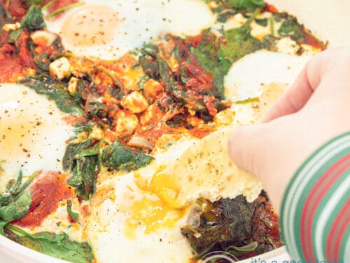Een vierkante foto met shakshuka in een koekenpan. Een hand dipt in het gepocheerde ei met wat brood.