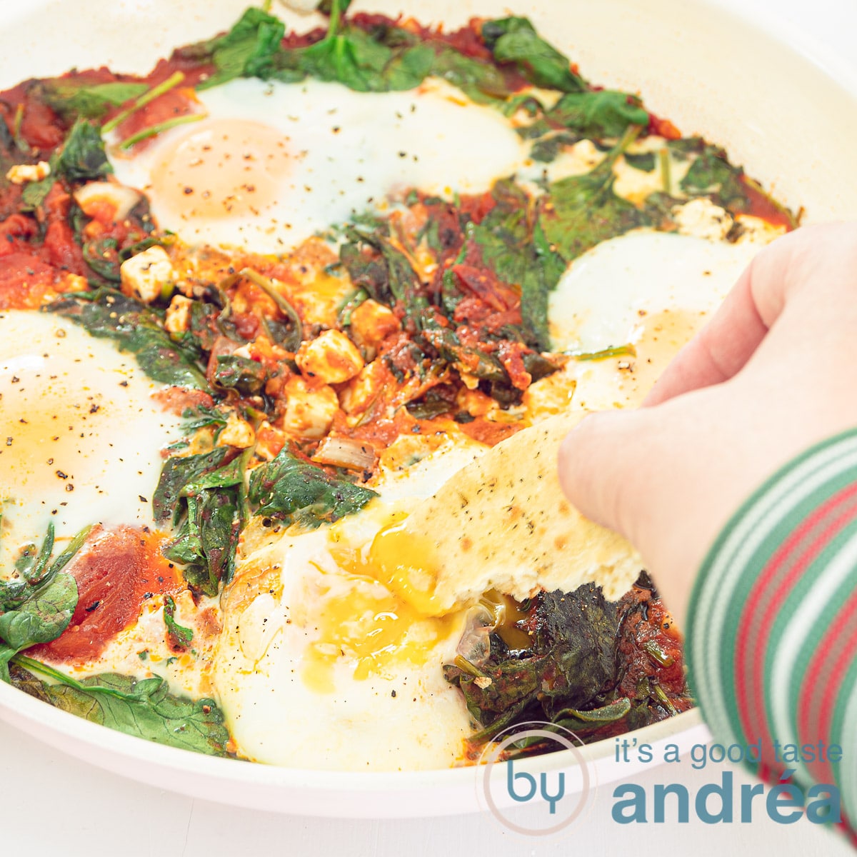 Een vierkante foto met shakshuka in een koekenpan. Een hand dipt in het gepocheerde ei met wat brood.