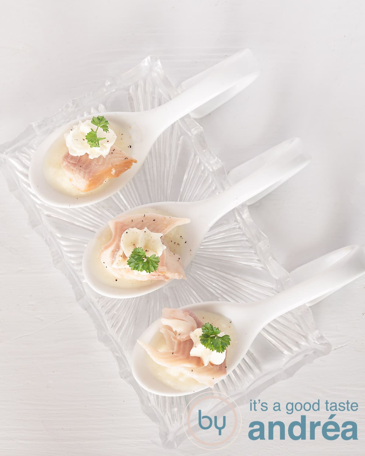 Een hoogte foto van drie amuselepels met aspergepuree, gerookte forel en mierikswortel mousse op een witte ondergrond