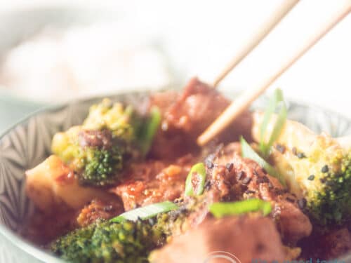 Een vierkante foto waar twee chopsticks een blokje biefstuk uit een groene kom gevuld met broccoli, biefstuk en saus pakt.