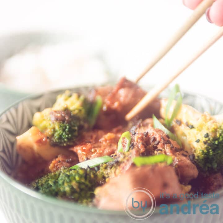 Een vierkante foto waar twee chopsticks een blokje biefstuk uit een groene kom gevuld met broccoli, biefstuk en saus pakt.