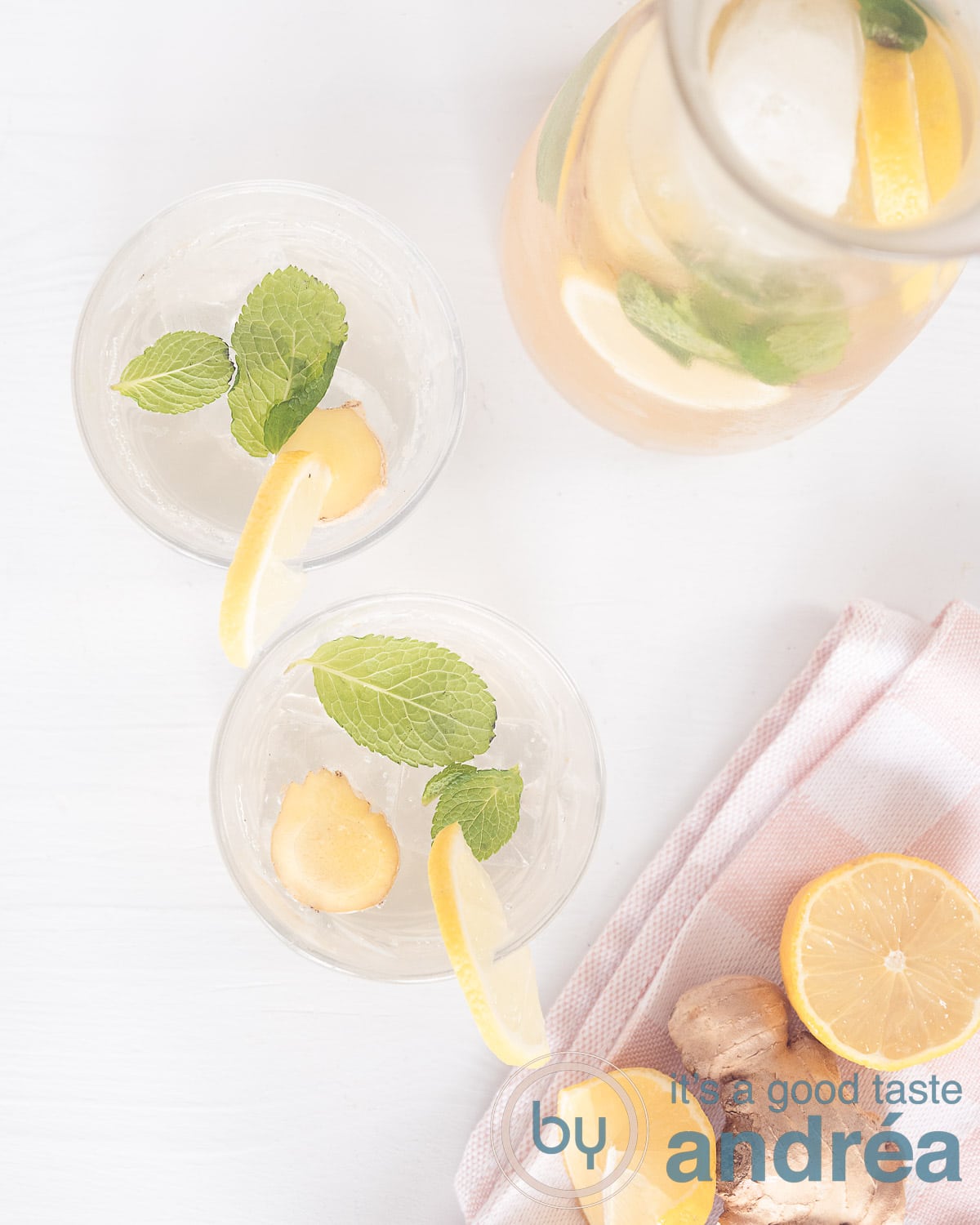 Een foto van bovenaf met twee glazen gevuld met citroen-gember limonade alsmede een karaf. Onderin een roze wite theedoek met citroen en een pers