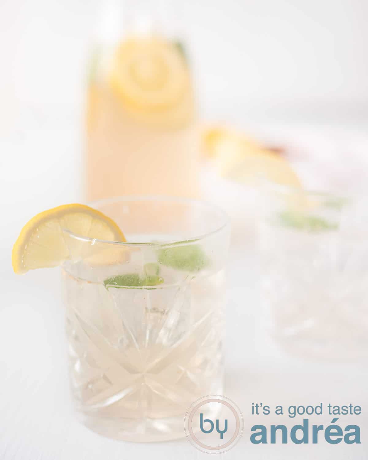 Een hoogtefoto met een glas gevuld met limonade met gember en citroen. Een karaf in de achtergrond alles op een witte ondergrond