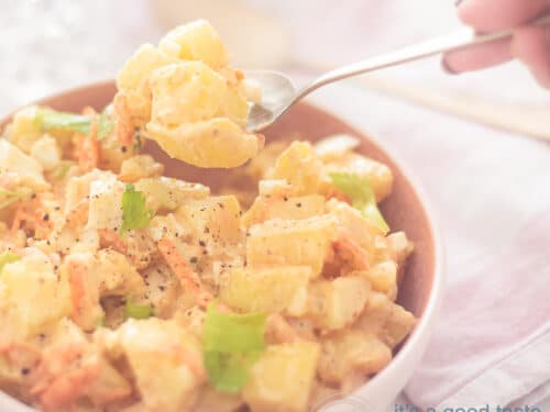 Een vierkante foto van een roze schaal met aardappelsalade. Een hand pakt een lepel eruit