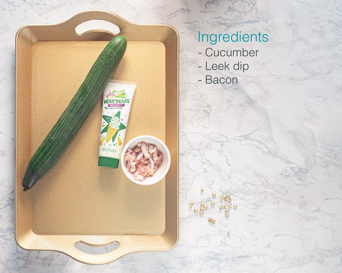Individually labeled ingredients on a golden tray to make cucumber appetizers with leek dip and bacon laid out on a white marble backdrop