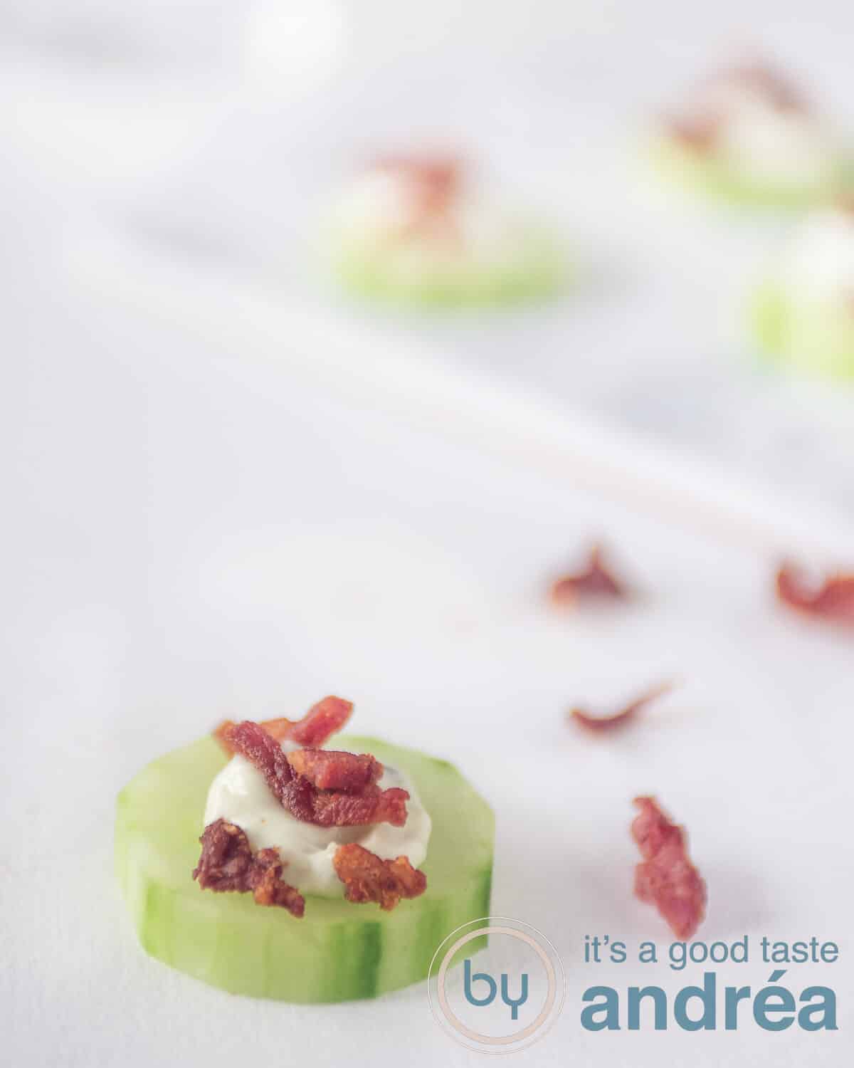 A cucumber appetizer topped with leek dip and bacon on a white background. In the back crisp bacon strips and more cucumber bite appetizers