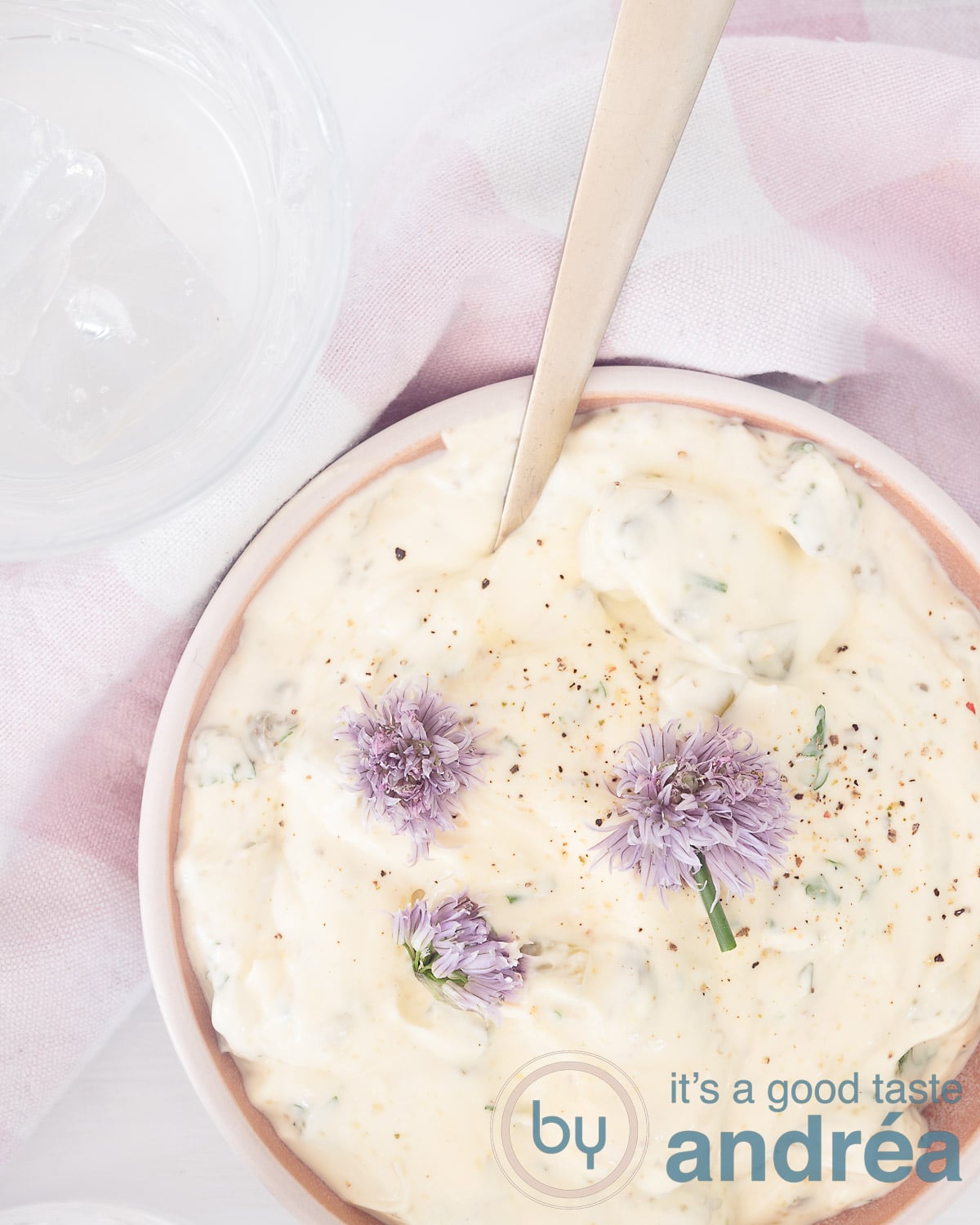 Een foto van bovenaf van een schaal gevuld met een sauce tartare, bieslookbloemetjes en een roze witte theedoek