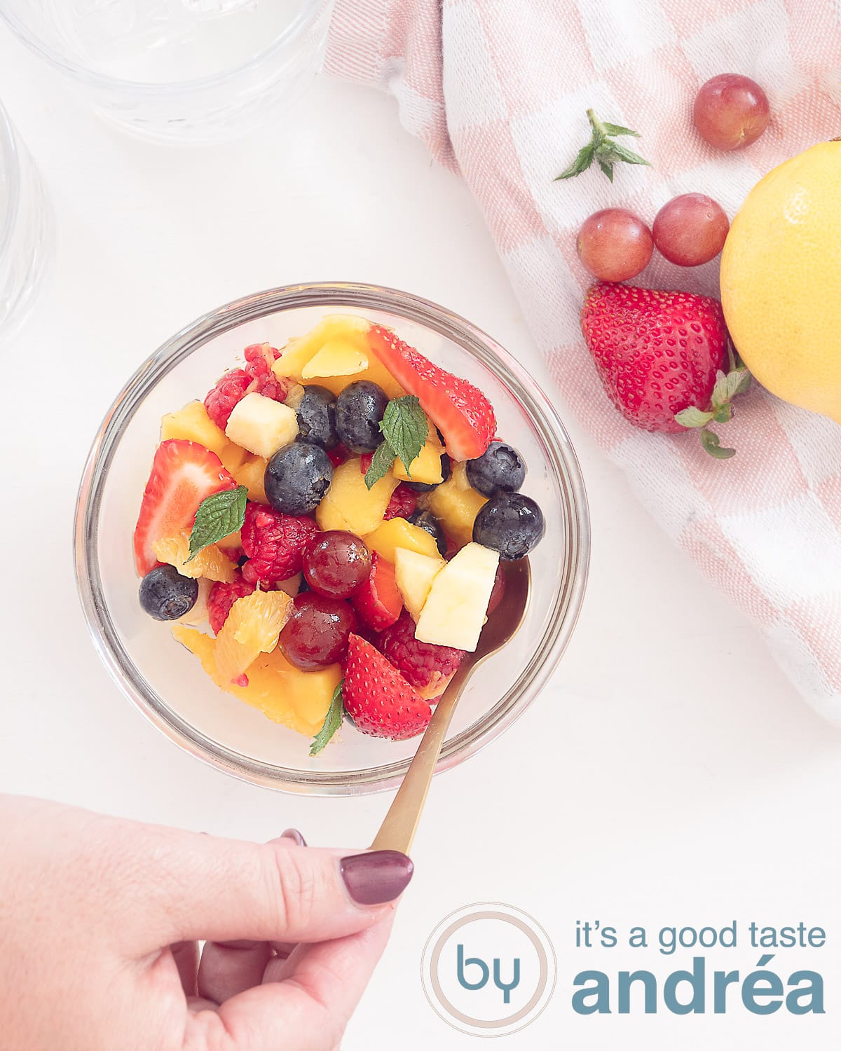 Een fruitsalade van bovenaf op een witte ondergrond, waar een hand een lepel van pakt. Rechts bovenin vers fruit op een roze witte doek