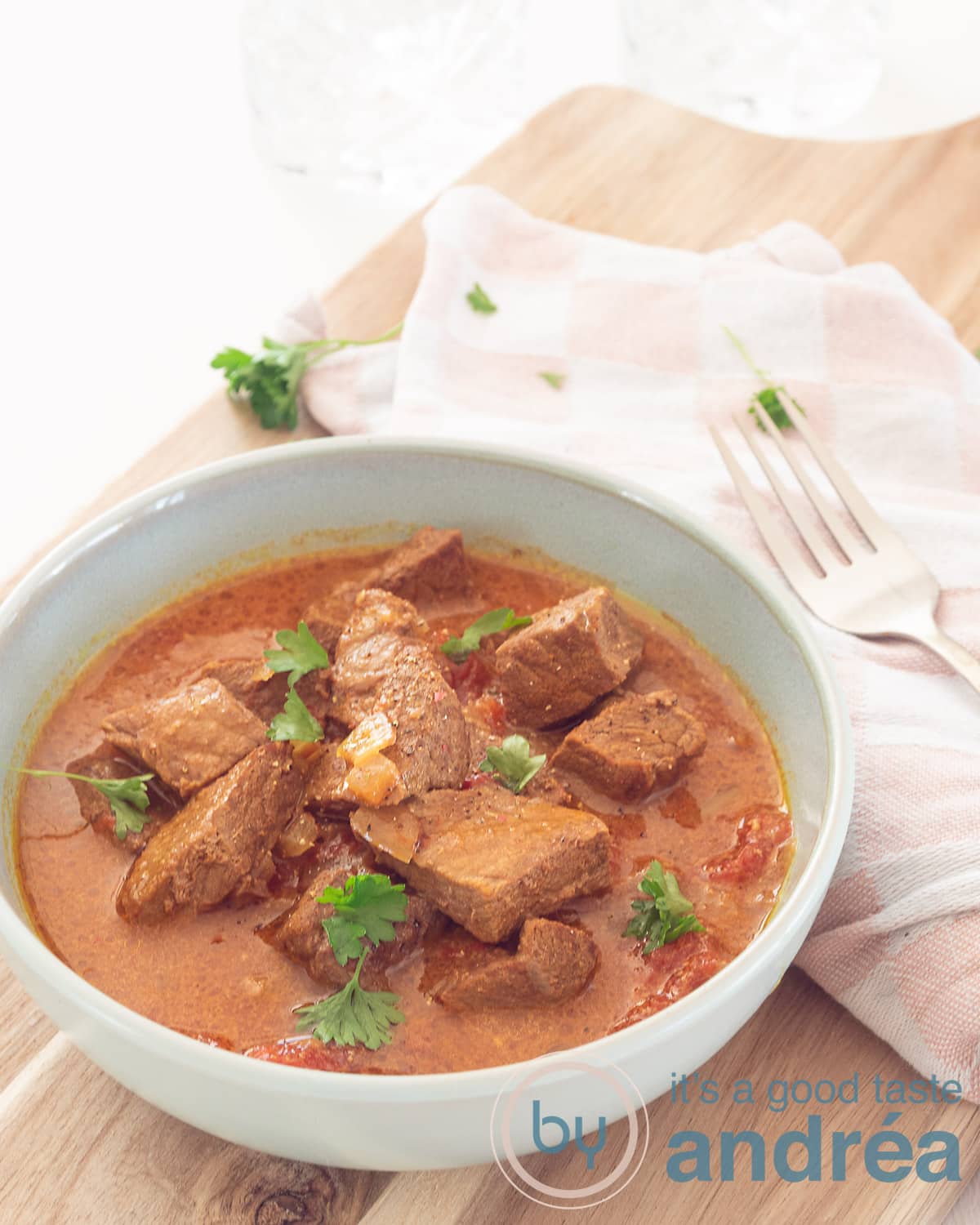 Een hoogtefoto met linksonderin een blauwe kom gevuld met bombay curry bestrooid met peterseile. Alles op een houten plank met een doek er schuin boven