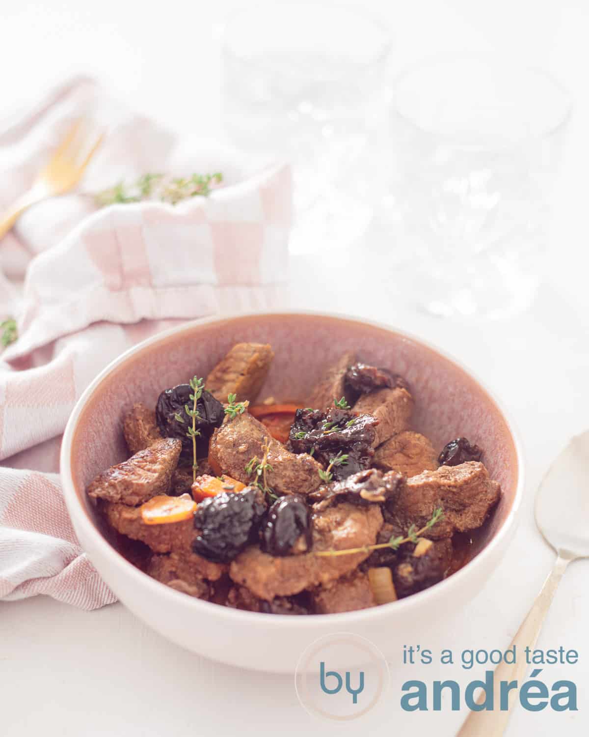 Een hoogtefoto met in het midden een slowcooker stoofpot met pruimen, rundvlees en wortel. Linksachter een roze witte theedoek, achterin twee glazen op een witte ondergrond. Rechts van de chaal een gouden lepel.