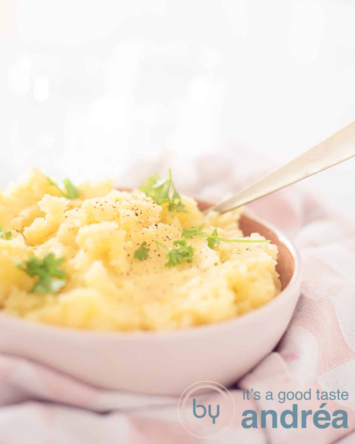 Een hoogtefoto met een deel van een roze schaal gevuld met aardappel puree gegarneerd met peterselie en op smaak gebracht met knoflook. Een gouden lepel steekt erin. Het staat op een roze witte theedoek.