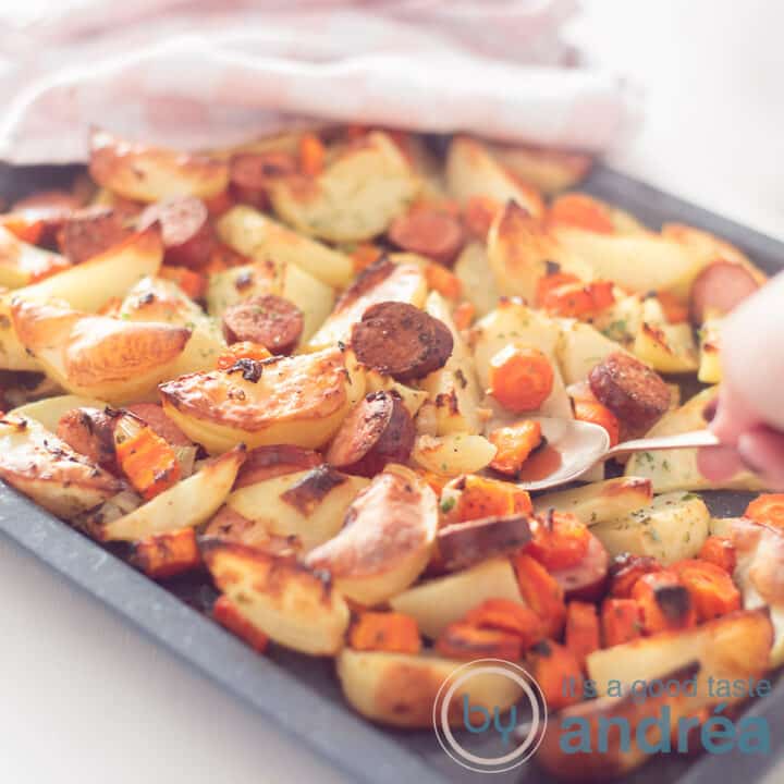 Een vierkante foto van een bakplaat gevuld met geroosterde aardappel, wortel en rookworst. Een lepel neemt een schep eruit. In de achtergrond een roze witte theedoek.