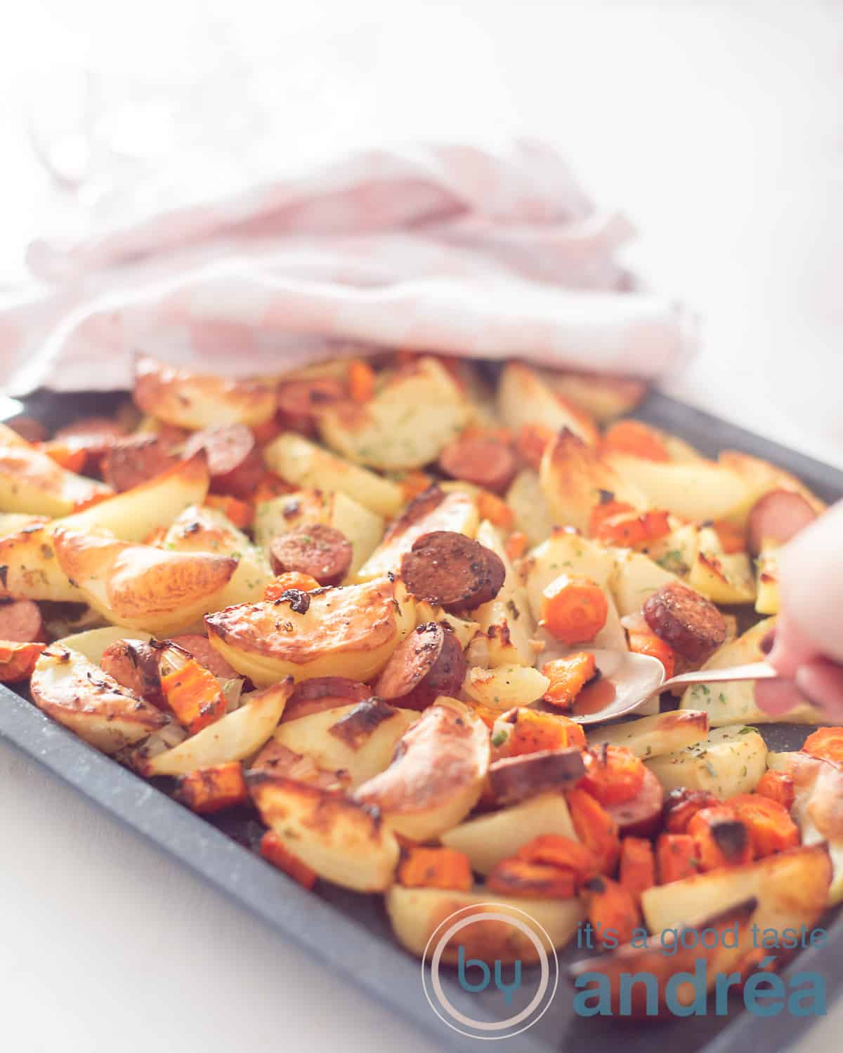 Een hoogtefoto met een traybake van aardappel wedges, wortel en rookworst. Een lepel schept eruit. In de achtergrond een roze witte theedoek.