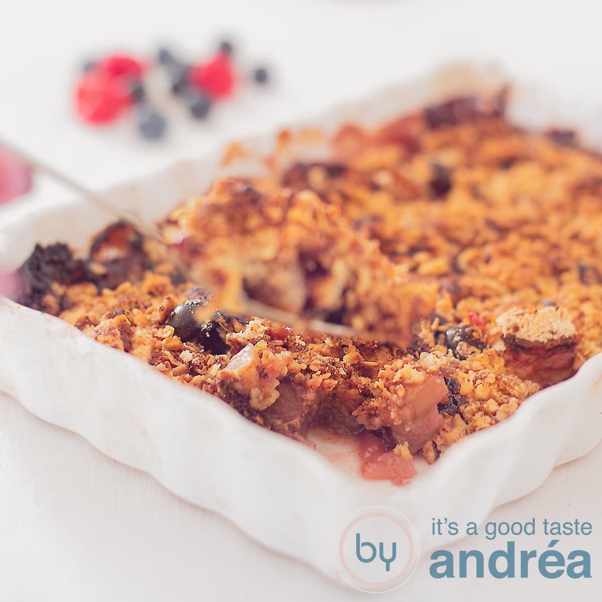 Een vierkante foto met een witte ovenschaal gevuld met frambozen havermout crumble. Een hand houdt een lepel vast met crumble. Een witte ondergrond met in de achtergrond bessen en frambozen