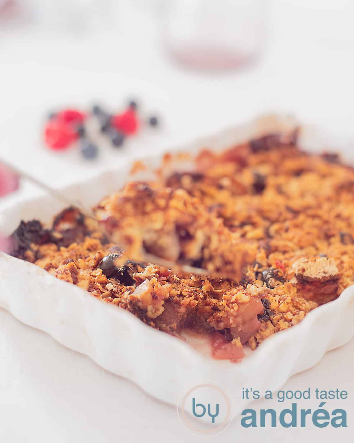 Een hoogtefoto met een witte ovenschaal gevuld met een appel bessen crisp op een witte ondergrond. In de achtergrond blauwe bessen en frambozen. Een hand houdt een lepel met crumble omhoog.