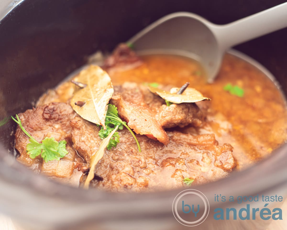 Een breedtefoto met een slowcooker gevuld met Stoofvlees, met laurierblad en peterselie. Een grijze opscheplepel neemt een beetje eruit.
