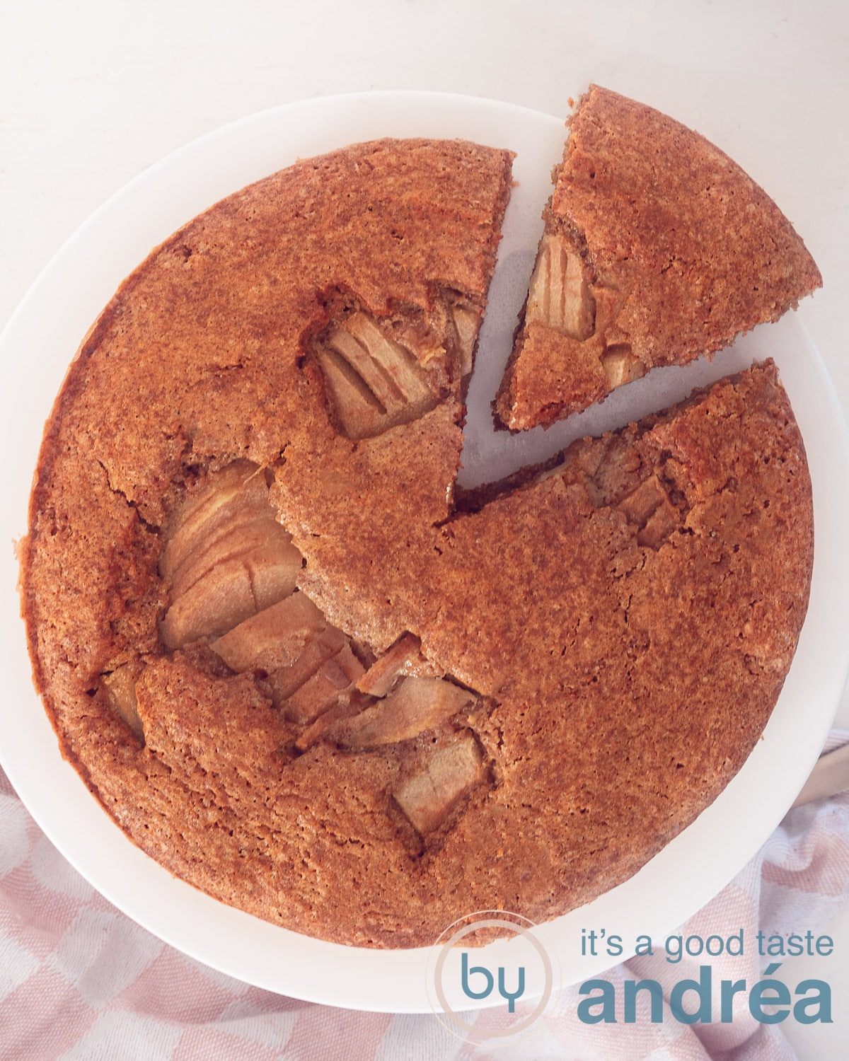 Een foto van bovenaf van een witte ondergrond een wit cakeplateau en een cake met peren waar een punt uit is gesneden die iets los ligt.