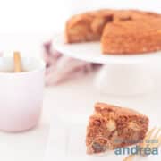 Een vierkante foto met een stukje perencake op de voorgrond en in de achtergrond een cake standaard met de rest van de cake. Links een roze kopje met koffie.