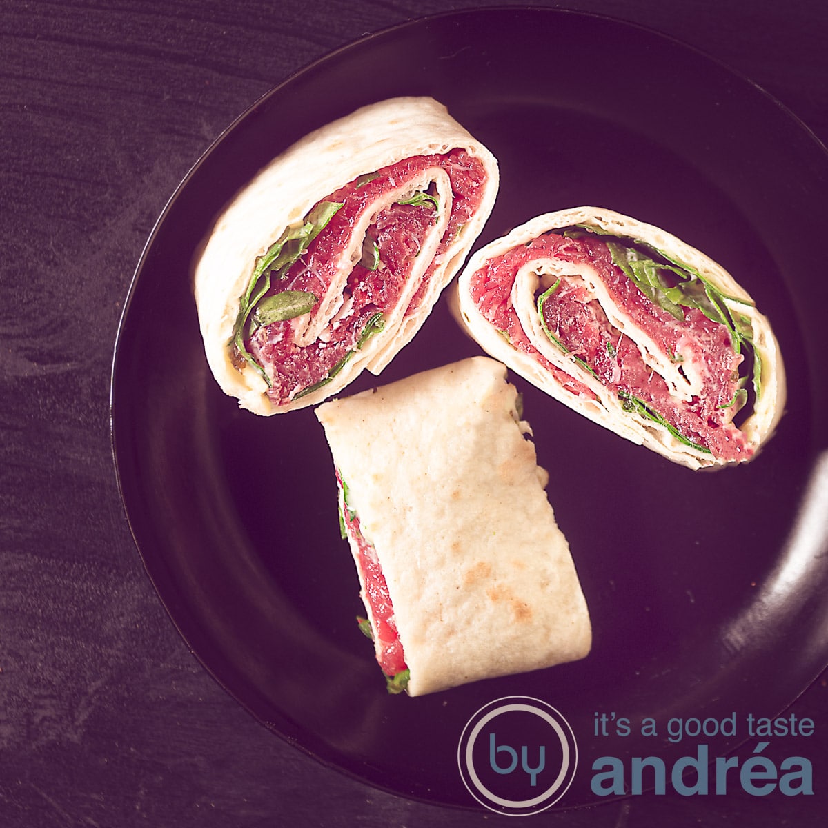 Een vierkante foto van een zwarte ondergrond met een zwart borjde met drie tortilla hapjes gevuld met carpacio, rucola, parmezaanse kaas.