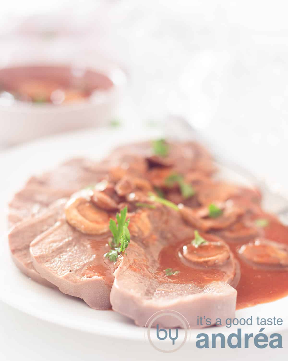 Een hoogtefoto met een witte schaal met plakjes rundertong overgoten met madeirasaus. Een witte achtergrond met een schaaltje met extra saus