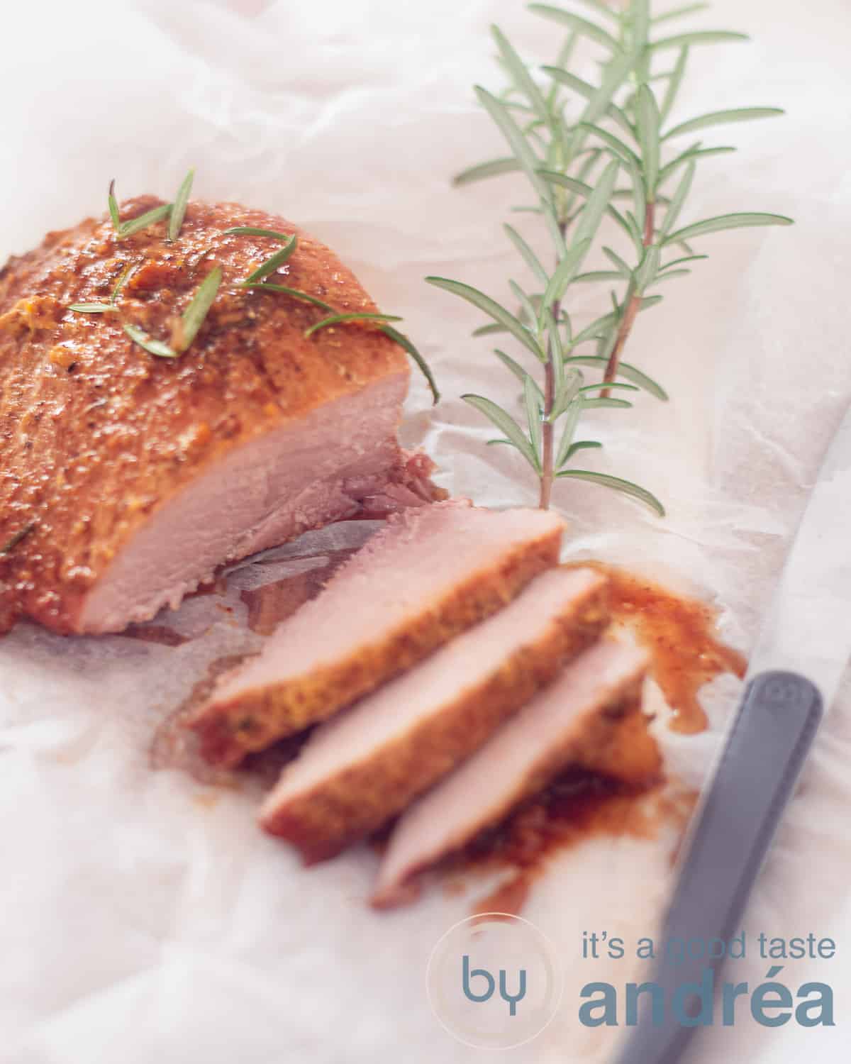 Een hoogtefoto met een ham met maple glaze waarvan drie plakjes zijn afgesneden. Liggend op een wit bakpapier, aan de rechterkant een steak mes en bovenin rozemarijn takje
