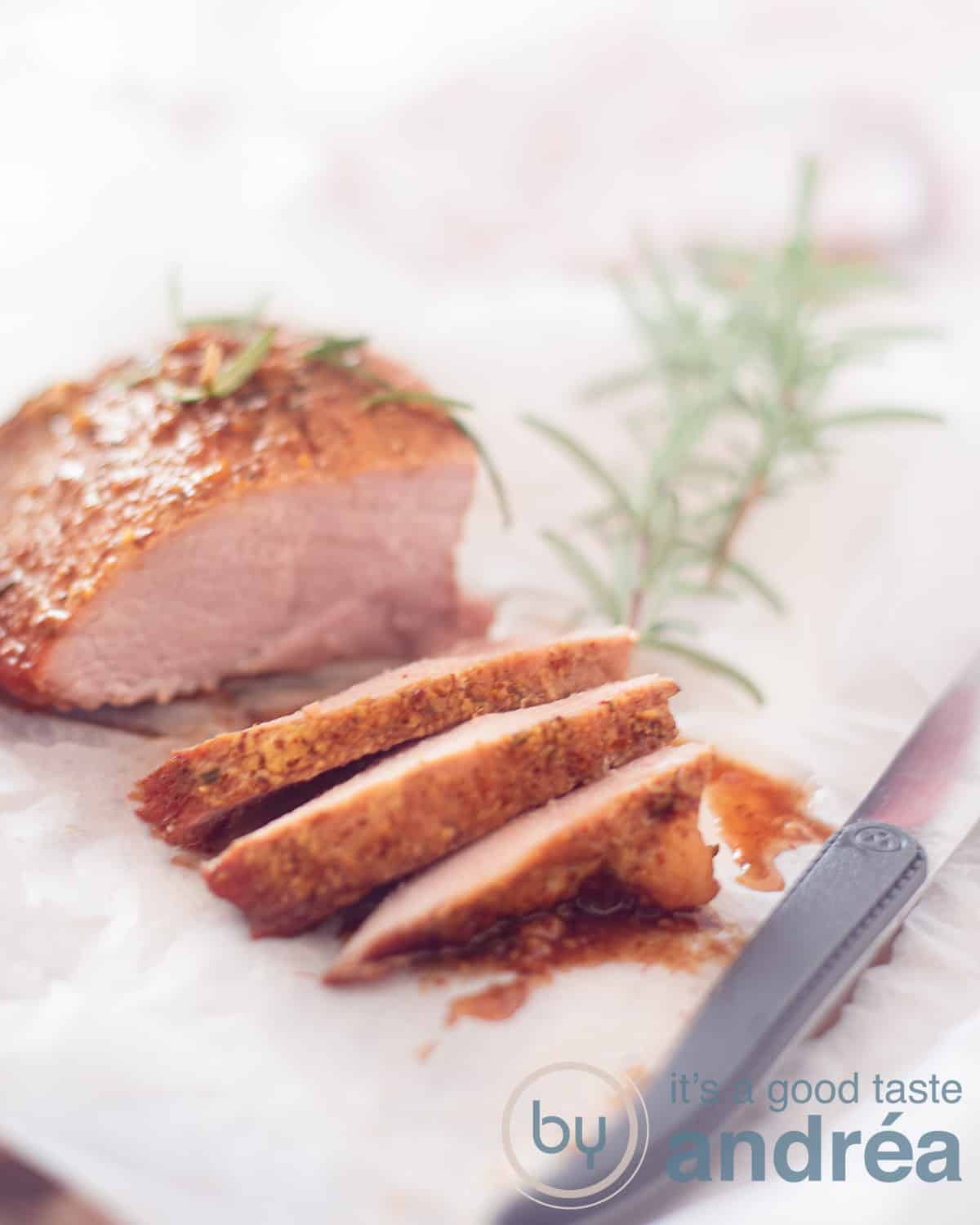 Een ham op een wit bakpapier, drie plakjes eraf gesneden. Een takje rozemarijn rechts erboven. Rechts onderin een steak mes