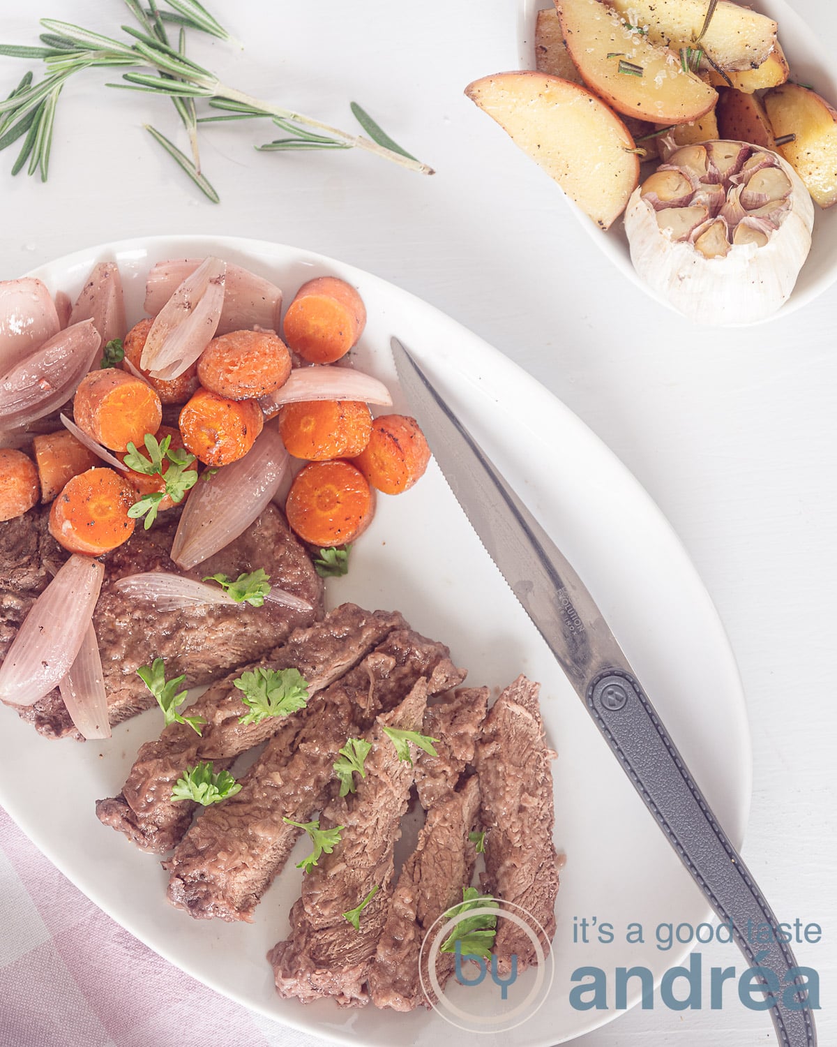 Een foto van bovenaf, rechtsonderin een witte schaal met een steak mes, reepjes entrecote, worteltjes en sjalotjes. Links bovenin aardappeltjes met knoflook.