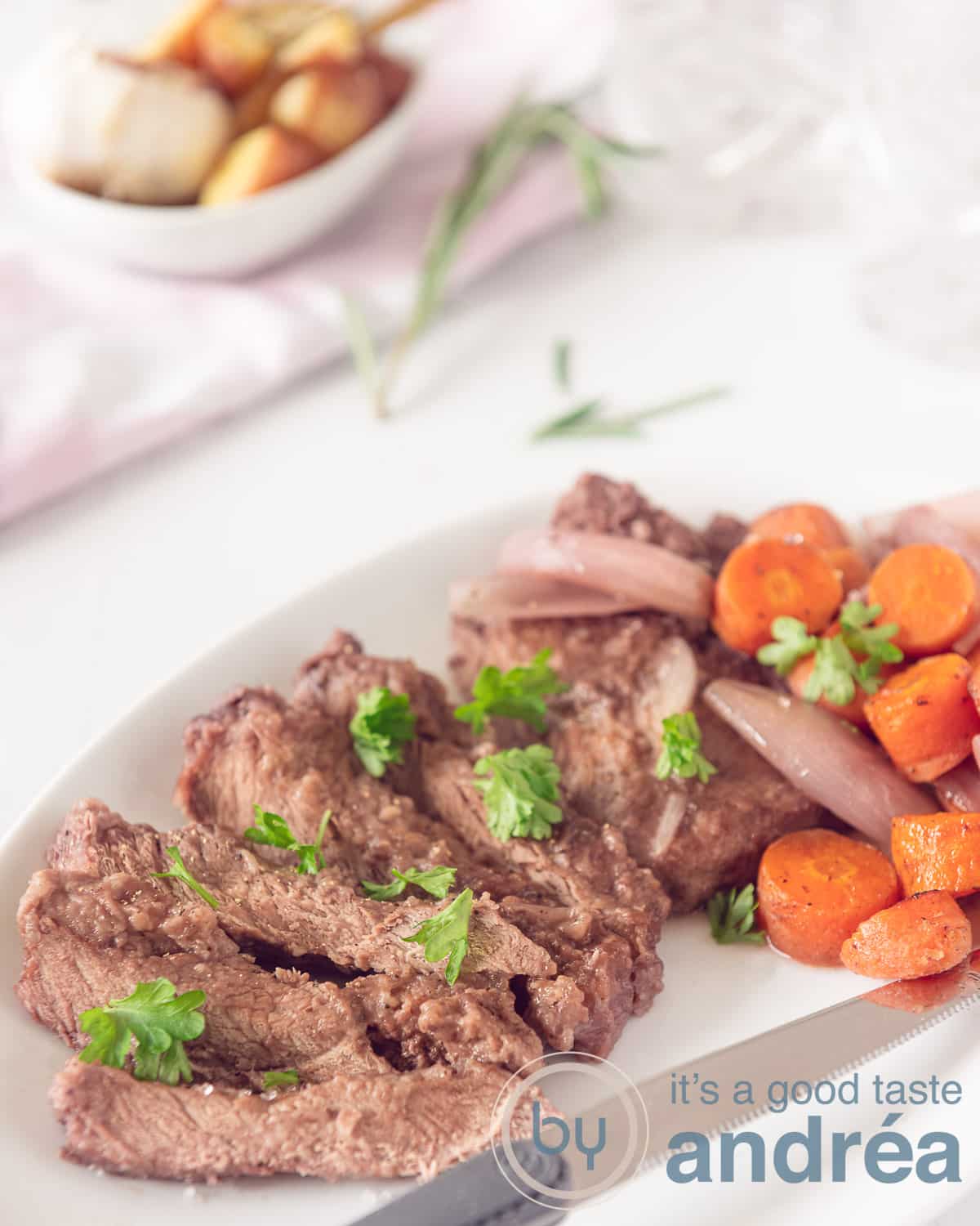 Onderin een schaal met in wijn gestoofde entrecote, sjalot en worteltjes. Bovenin een schaal geroosterde aardappels met knoflook op een witte ondergrond.