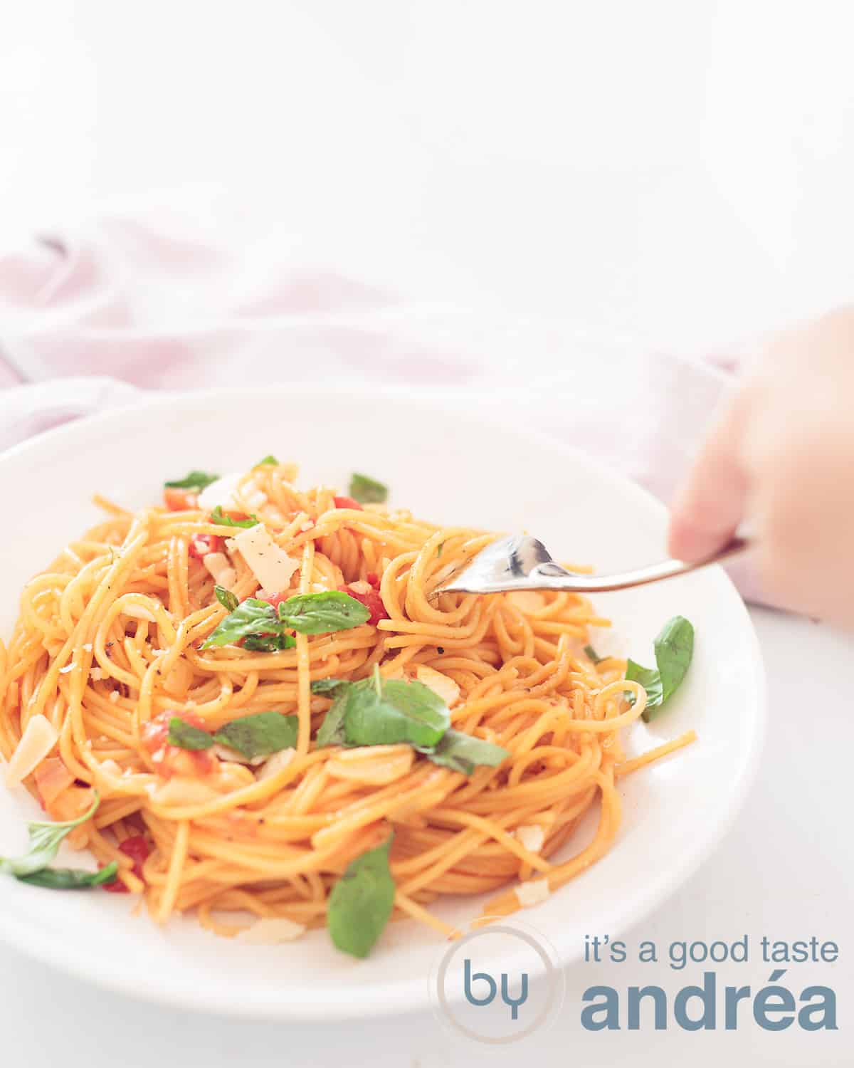 Een hoogtefoto met linksonder een wit bord gevuld met pasta, kaas, tomaat en basilicum. Een vork prikt een hapje. een witte ondergrond. Linksachterin een roze witte theeodek
