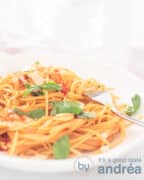Een hoogtefoto met een deel van een diep wit bord met spaghetti, basilicum, kaas, en tomatensaus. Een gouden vork prikt erin. Een witte ondergrond.