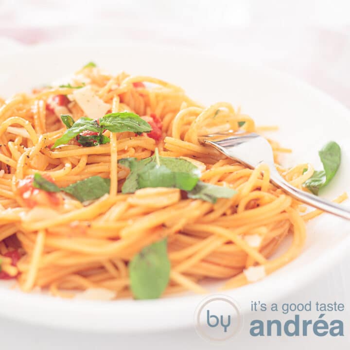 Een vierkante foto met een wit bord gevuld met spaghetti, kaas, tomaat en bestrooid met basilicumblaadjes. Een gouden vork steekt erin.