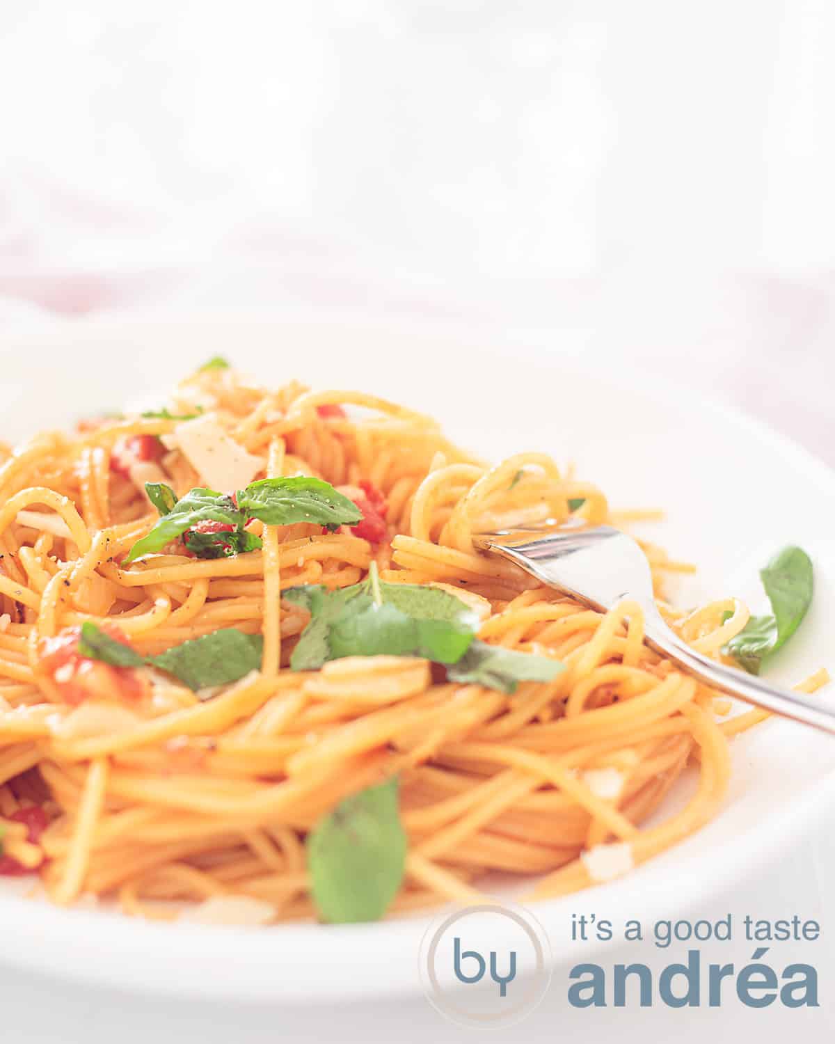 Een hoogtefoto met een deel van een diep wit bord met spaghetti, basilicum, kaas, en tomatensaus. Een gouden vork prikt erin. Een witte ondergrond.