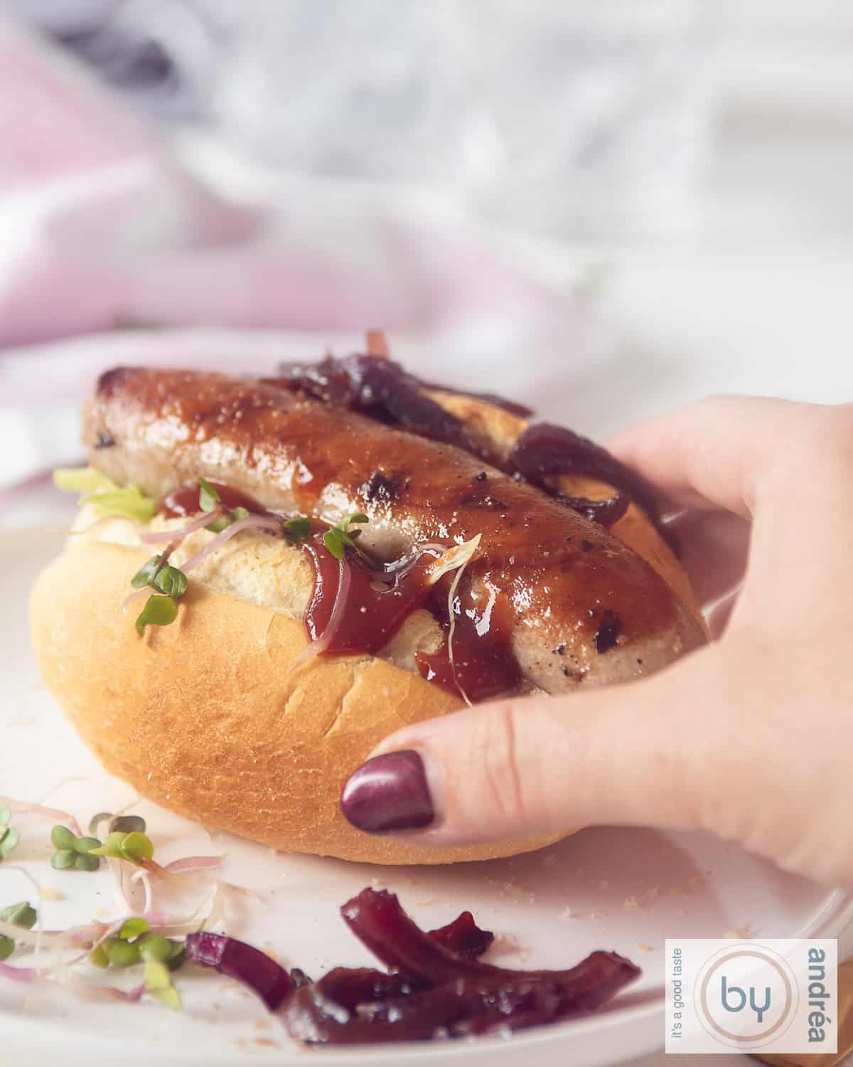 Een hand pakt een broodje met gegrilde braadworst, curry en uitjes van een wit bord