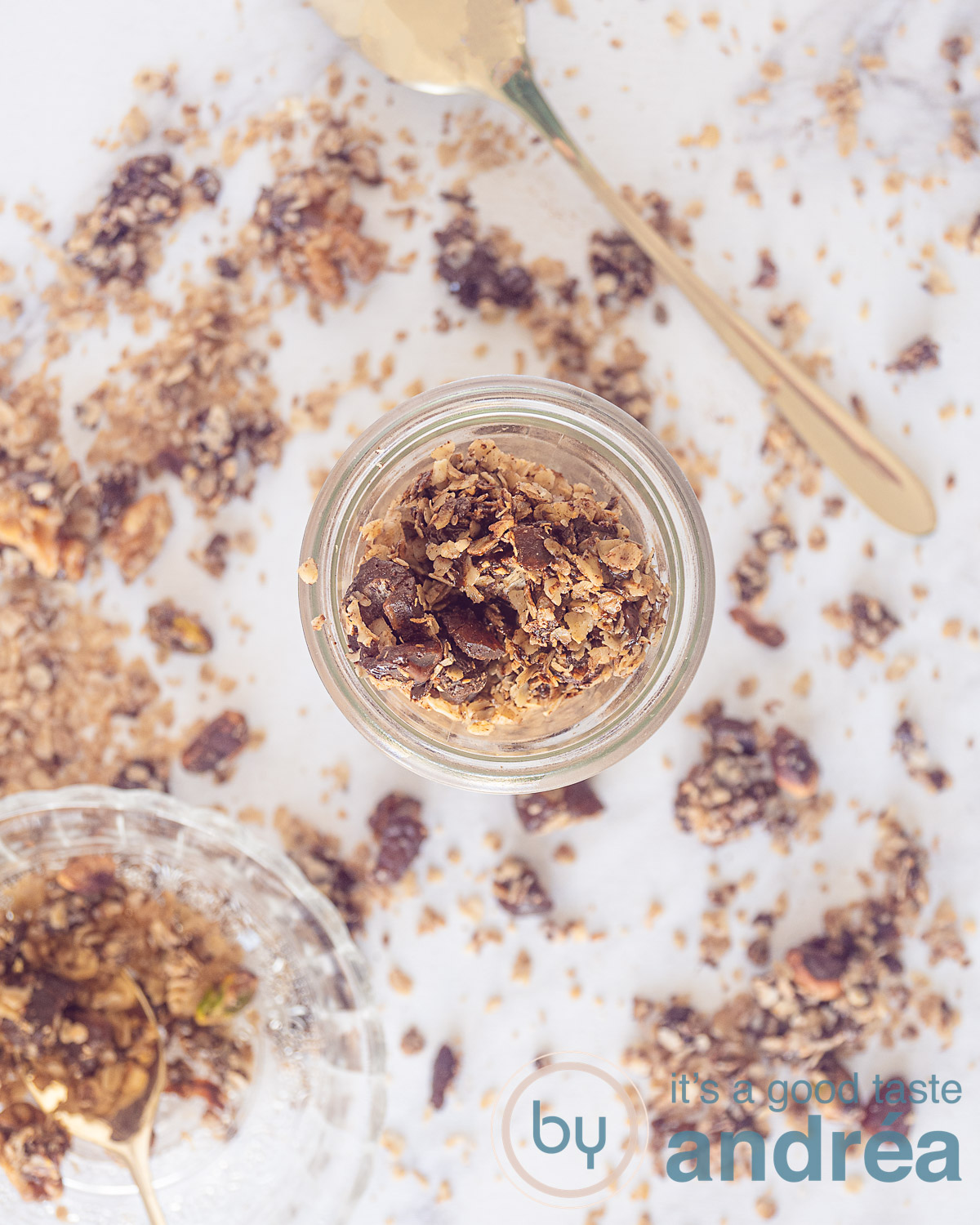 Een foto van bovenaf met een glazen flesje met daaromheen granola. Onderin een bakje met granola. Wat chocolade noten granola ligt eromheen verspreidt. Een gouden lepel rechts bovenin.