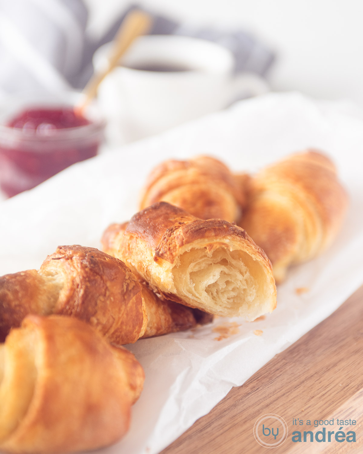 Croissants, zelfgemaakt op een bakpapier op een houten plank. Een croissant is doormidden, waardoor je de luchtige structuur goed kunt zien.