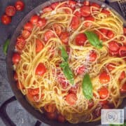 Een vierkante foto met een zwarte pan met linguine pasta, cherry tomaten, basilicum, ui en kaas op een grijze ondergrond.