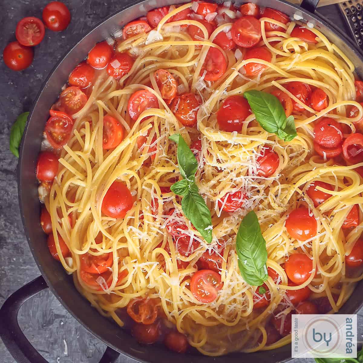 Een vierkante foto met een zwarte pan met linguine pasta, cherry tomaten, basilicum, ui en kaas op een grijze ondergrond.