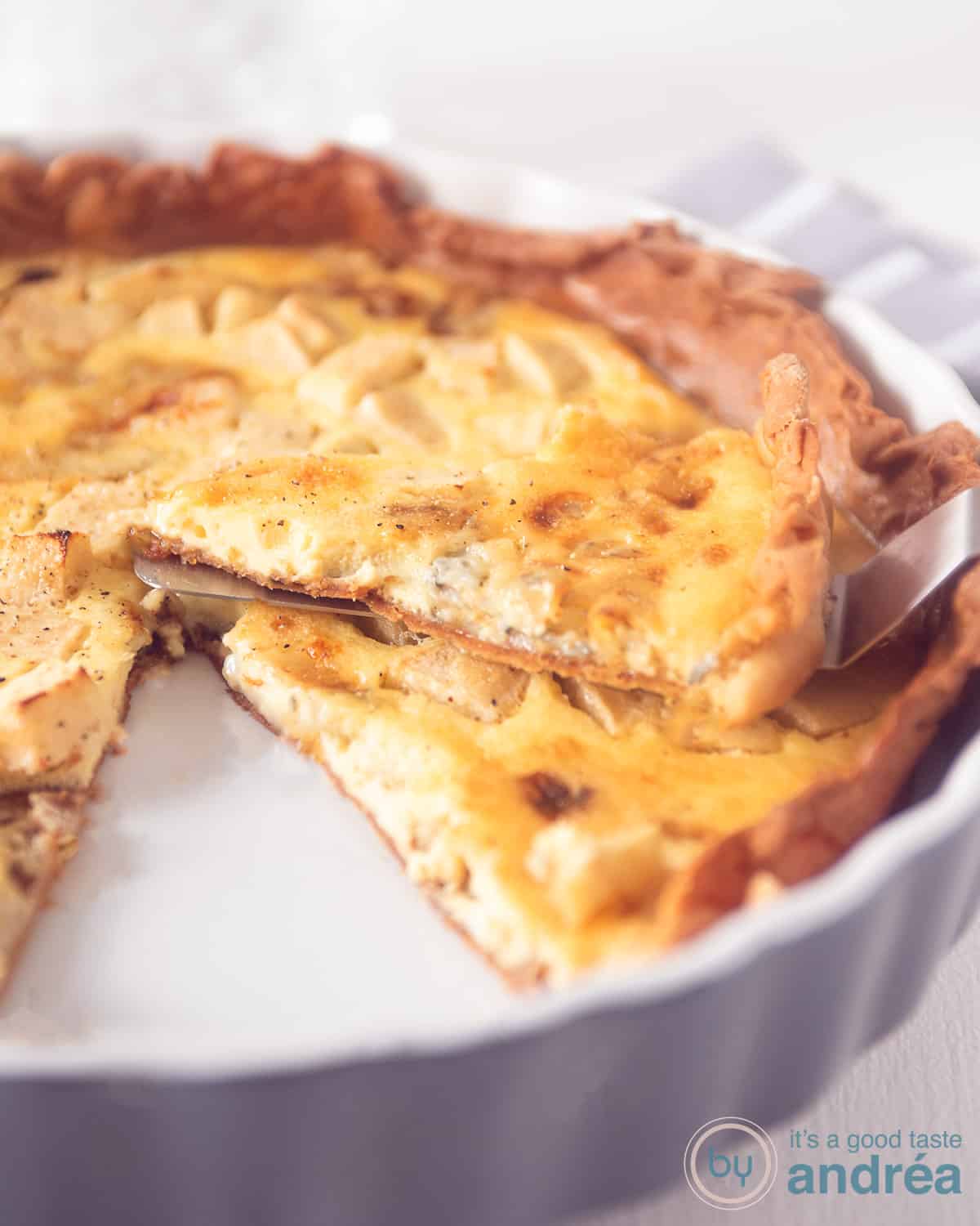 Een deel van een grijze quiche schaal met een gorgonzola appel quiche. Een part is eruit gesneden en ligt er bovenop.