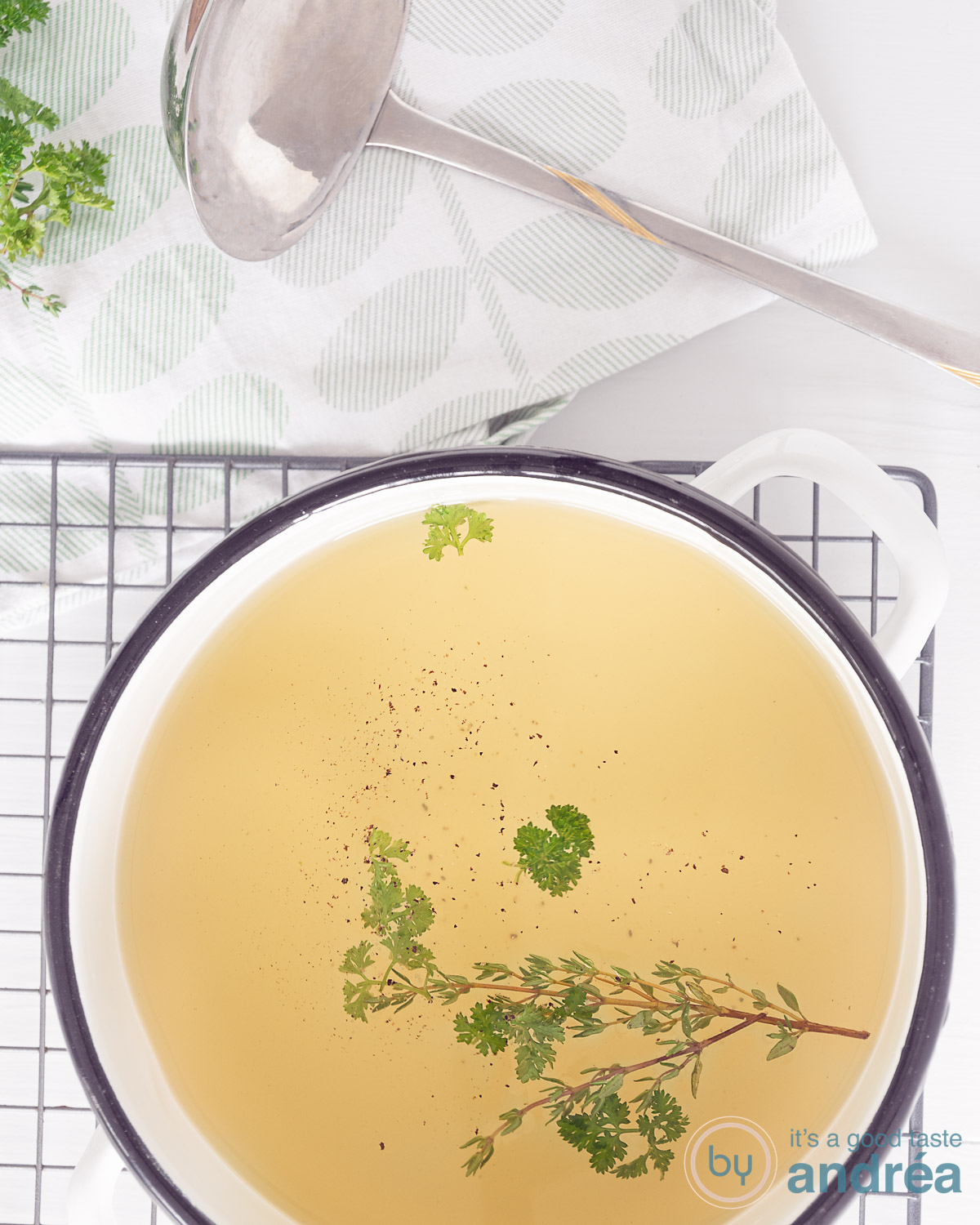 Groentebouillon in een soeppan op een rek. Bovenin een soeplepel en verse kruiden