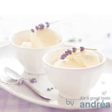 Twee witte kommetjes met lavendel ijs, een takje erbovenop. Op een wit schaaltje met een lepel aan de linkerkant. Staand op een geblokt tafelkleed