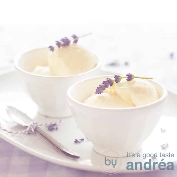Twee witte kommetjes met lavendel ijs, een takje erbovenop. Op een wit schaaltje met een lepel aan de linkerkant. Staand op een geblokt tafelkleed