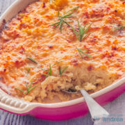 Een vierkante foto met een deel van een roze ovenschaal gevuld met macaroni en kaas, getopt met toefjes rozemarijn. Een lepel steekt erin. Een grijze ondergrond.