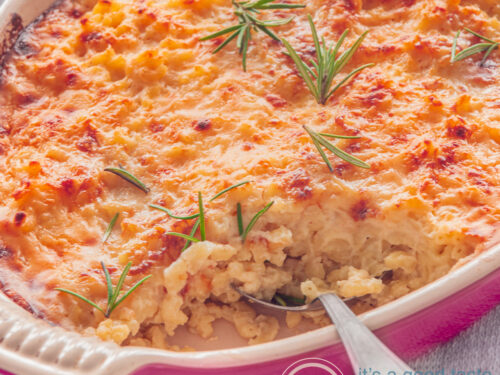 Een vierkante foto met een deel van een roze ovenschaal gevuld met macaroni en kaas, getopt met toefjes rozemarijn. Een lepel steekt erin. Een grijze ondergrond.