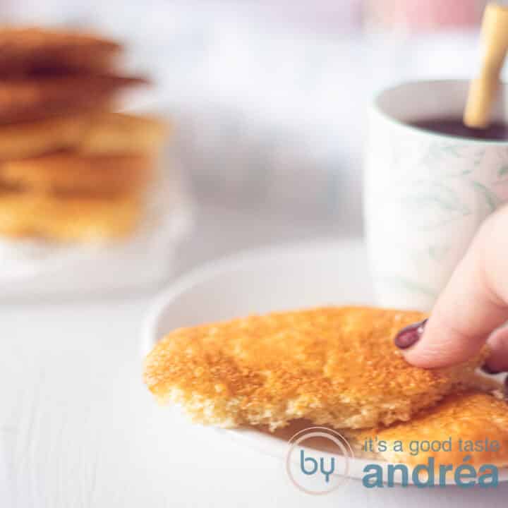 Een witte ondergrond met aan de rechterkant een wit bord met twee halve eierkoeken. Een hand pakt een eierkoek. Links achterin een stapel eierkoeken.