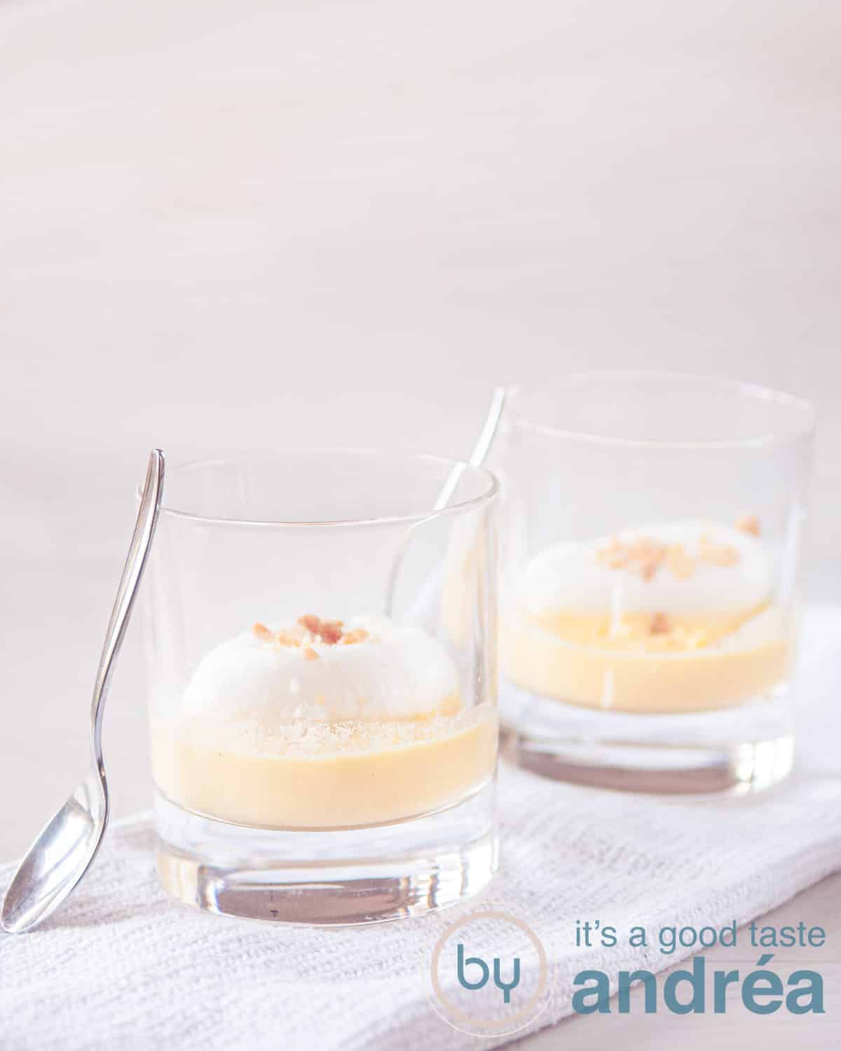 Twee glazen met Oeufs a la neige op een wit doek op een witte ondergrond. Twee lepels staan tegen de glazen