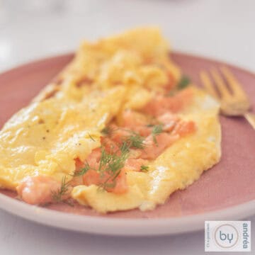 Een vierkante foto met een omelet gevuld met gerookte zalm en bieslook op een roze bord.