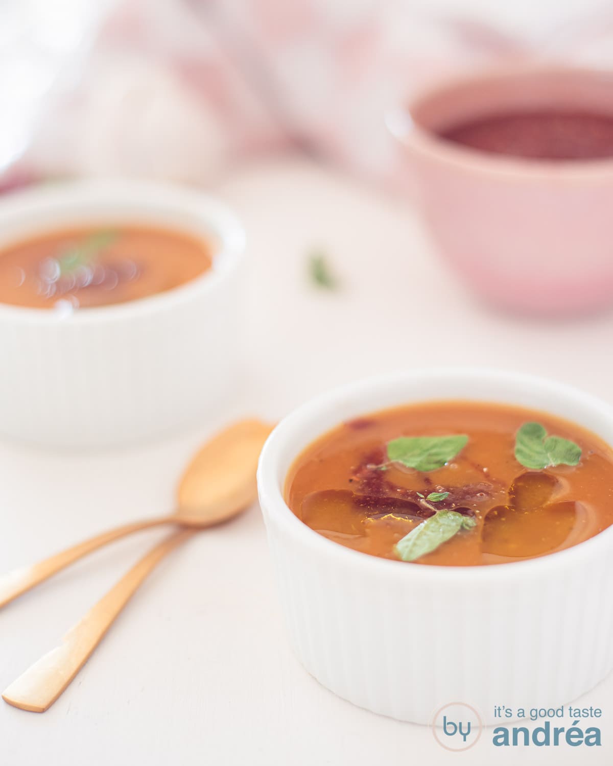 Pompoensoep met een pittige salsa in twee kommetjes op een witte ondergrond. Twee gouden lepels er links naast.