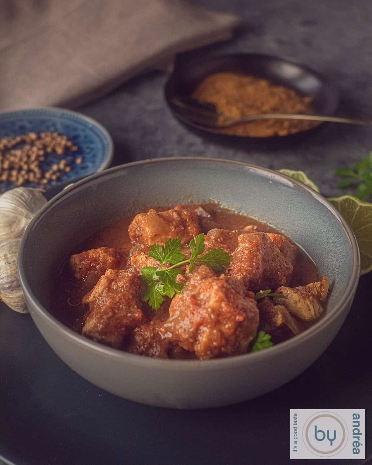 Een foto van bovenaf van een roze schaal met kip, curry saus en peterselie. Een lepel zit in de schaal. Een witte ondergrond met een roze witte theedoek rechts bovenin.