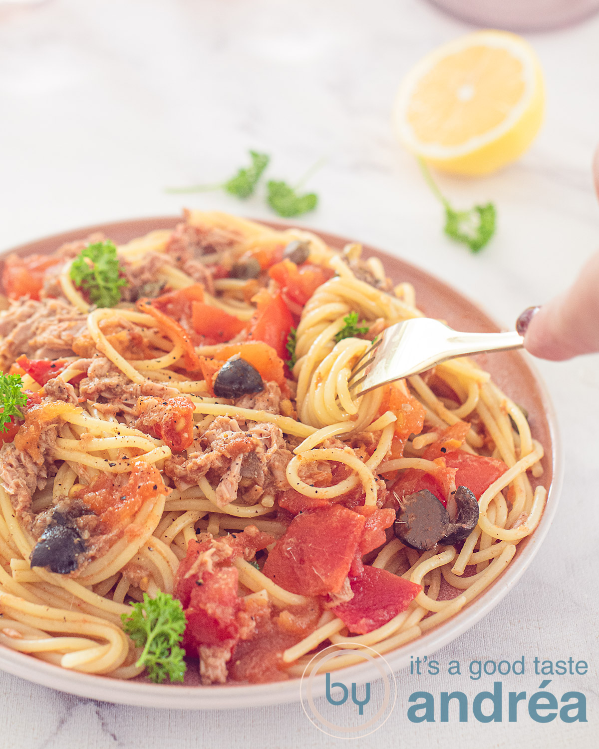 Een hoogtefoto met een roze bord met Spaghetti alla Puttanesca. Een vork heeft spaghetti eromheen gedraaid. Een roze witte theedoek, verse basilicum en glazen op een witte achtergrond.