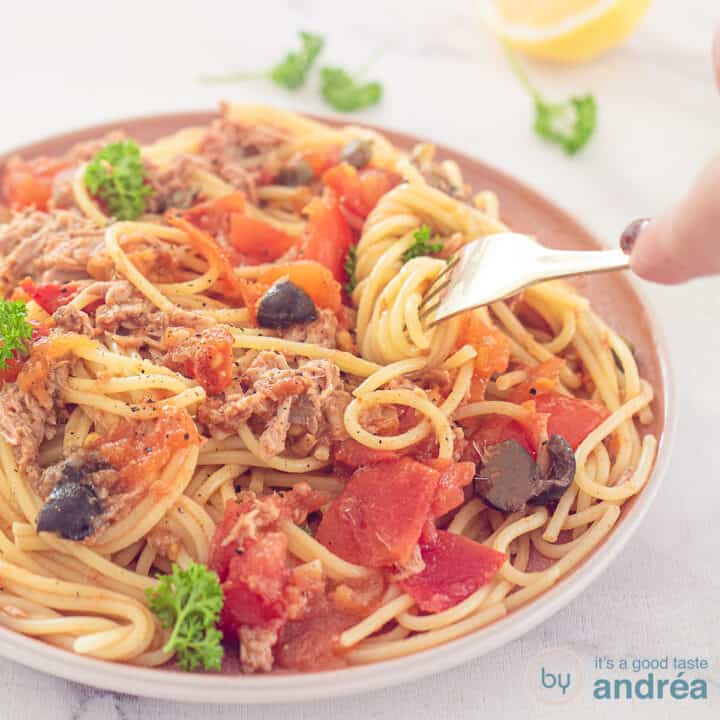 Een vierkante foto met een roze bord met daarop spaghetti Puttanesca. Spaghetti om een vork gedraaid. Een roze witte theedoek in de achtergrond.